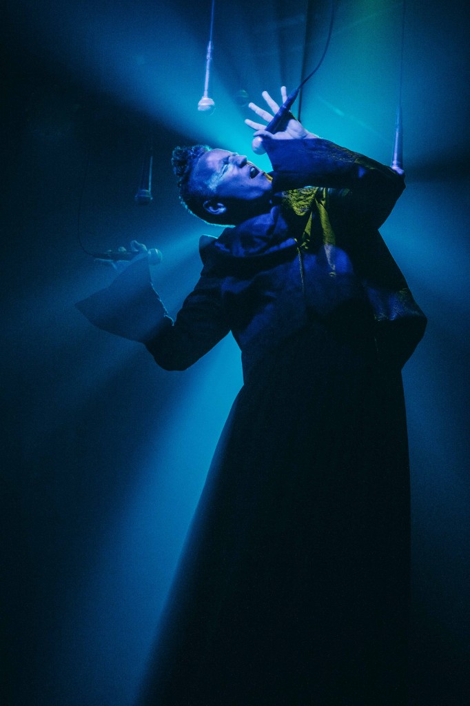 Stewart Legere performing in Let's Not Beat Each Other to Death. Blue light hits him from behind as he sings into one of seven microphones hanging from the ceiling.